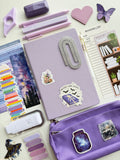 Purple stationery items including a notebook, pens, and stickers on a white background