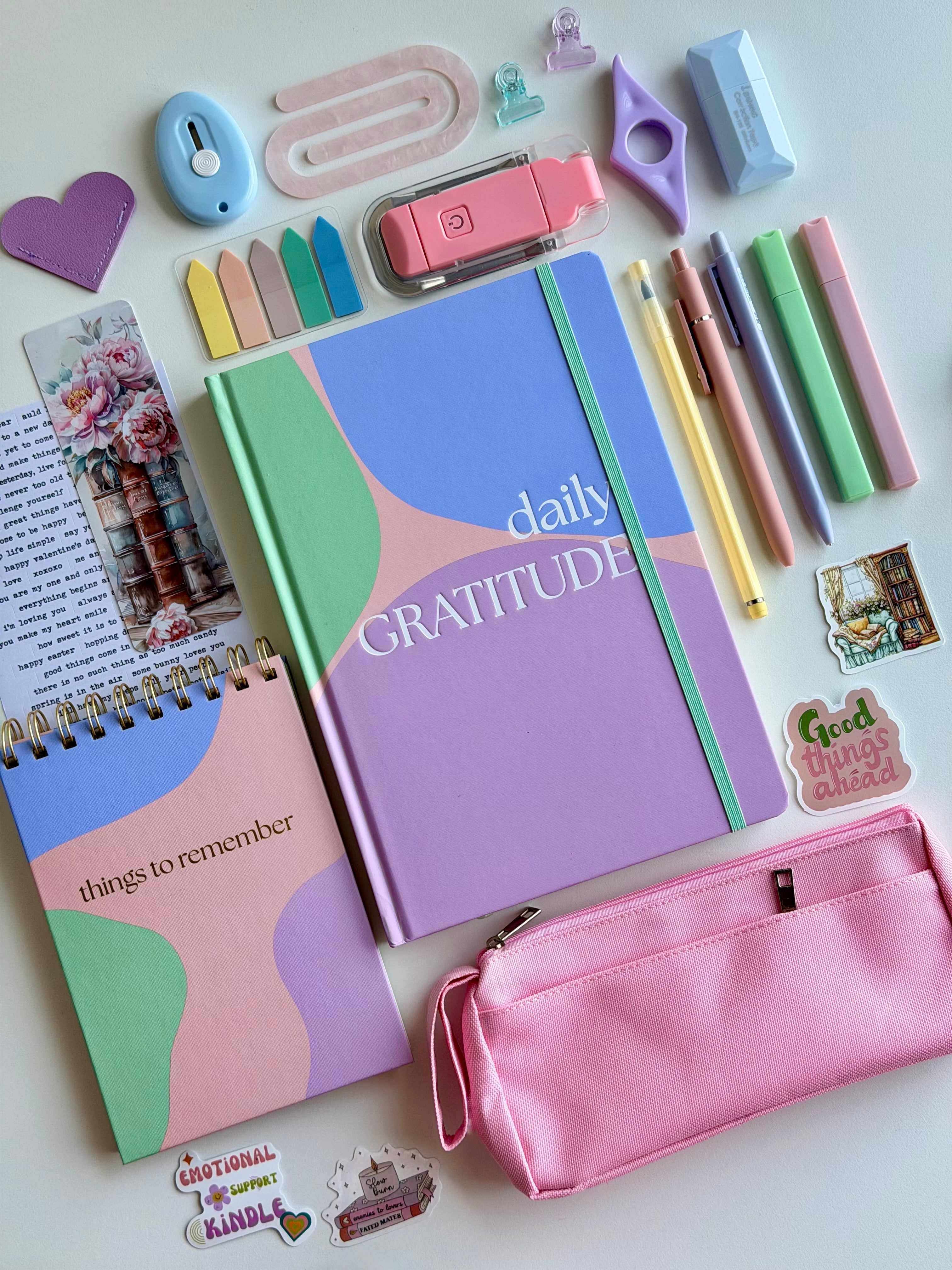 Stationery items including a gratitude journal, pink pencil case, and colorful markers on a white background.