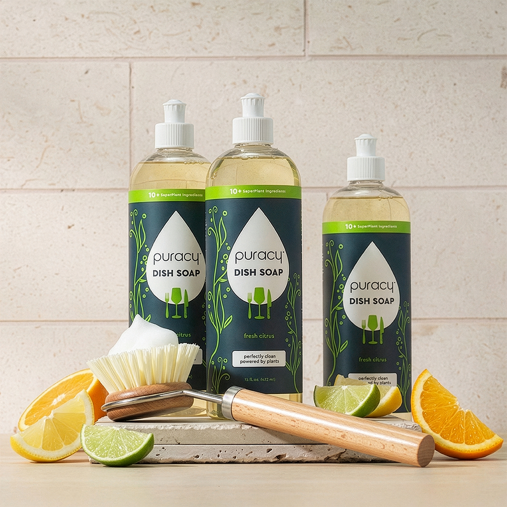 Three bottles of Puracy dish soap on a kitchen counter with a scrubber and citrus fruits.