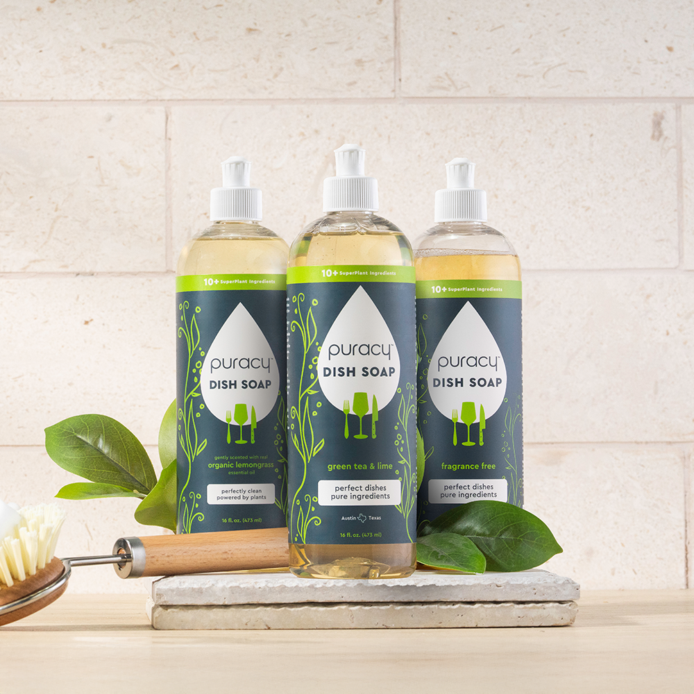 Three bottles of Puracy dish soap on a kitchen counter with a brush and leaves.