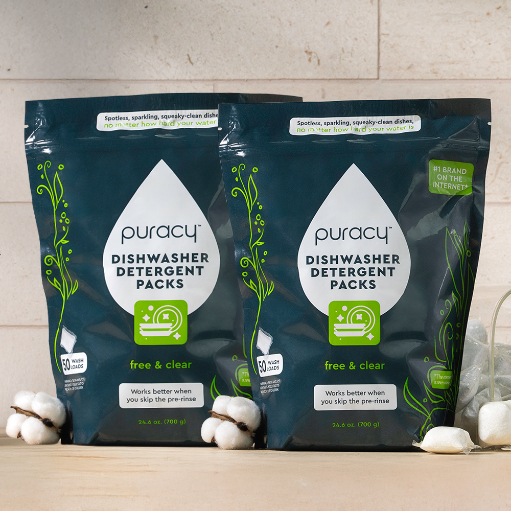 Two packages of Puracy Dishwasher Detergent Packs on a wooden surface with cotton balls.