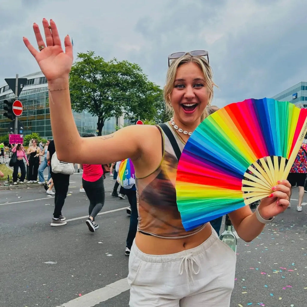 Person holding a rainbow-colored fan in an outdoor setting with people and buildings in the background