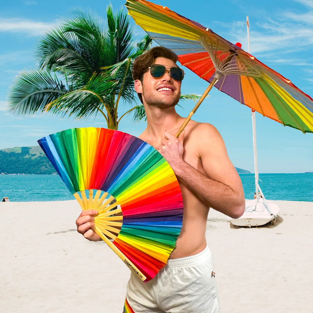 Man on a beach holding a rainbow-colored fan and umbrella with palm trees and ocean in the background.
