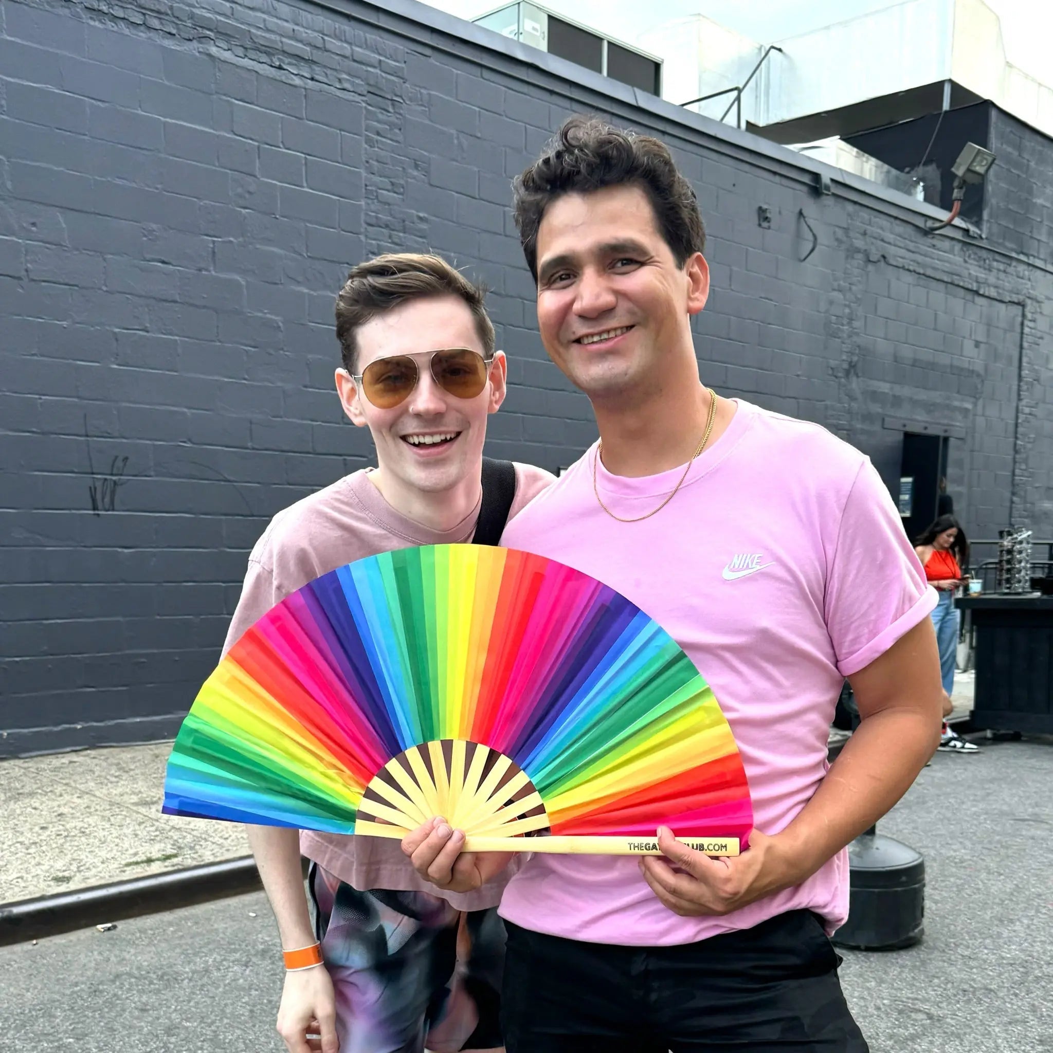 Two men standing outdoors, one holding a rainbow-colored fan, with a gray building in the background.
