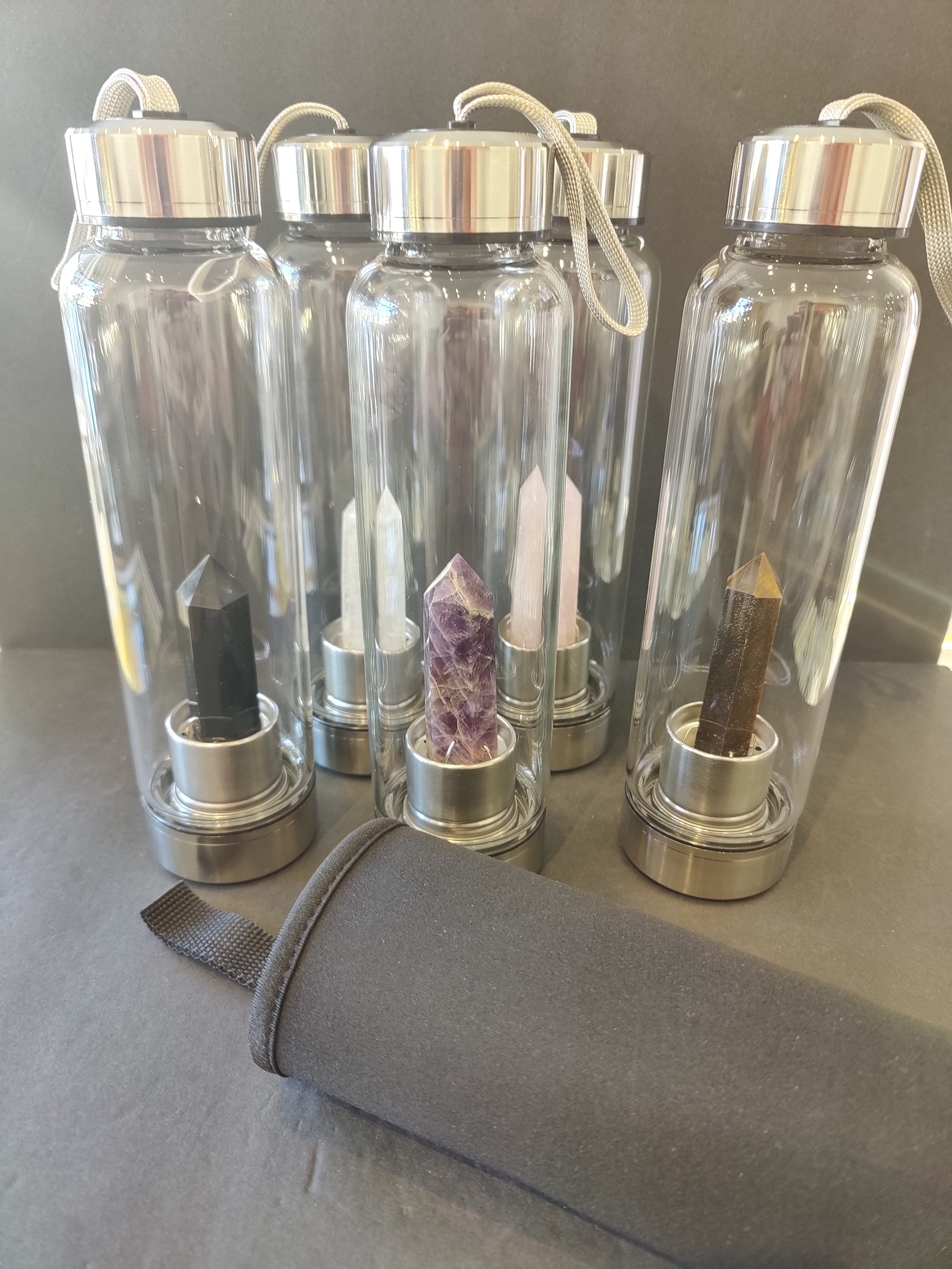 5 Clear water bottles 16oz with silver caps containing crystals and an insulated bottle cover on a gray surface.