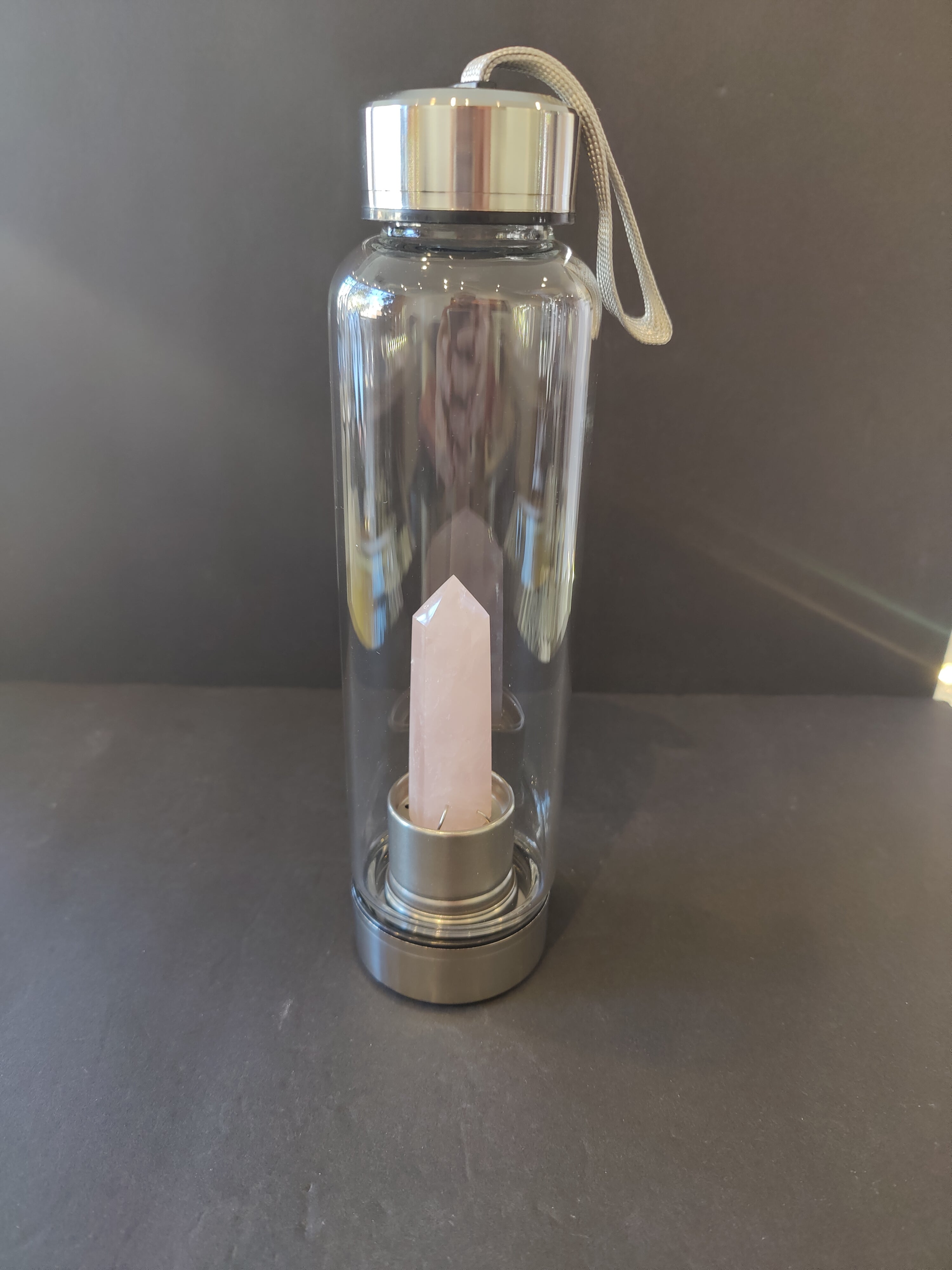 Clear glass water bottle with silver cap and rose quartz crystal inside on a dark surface