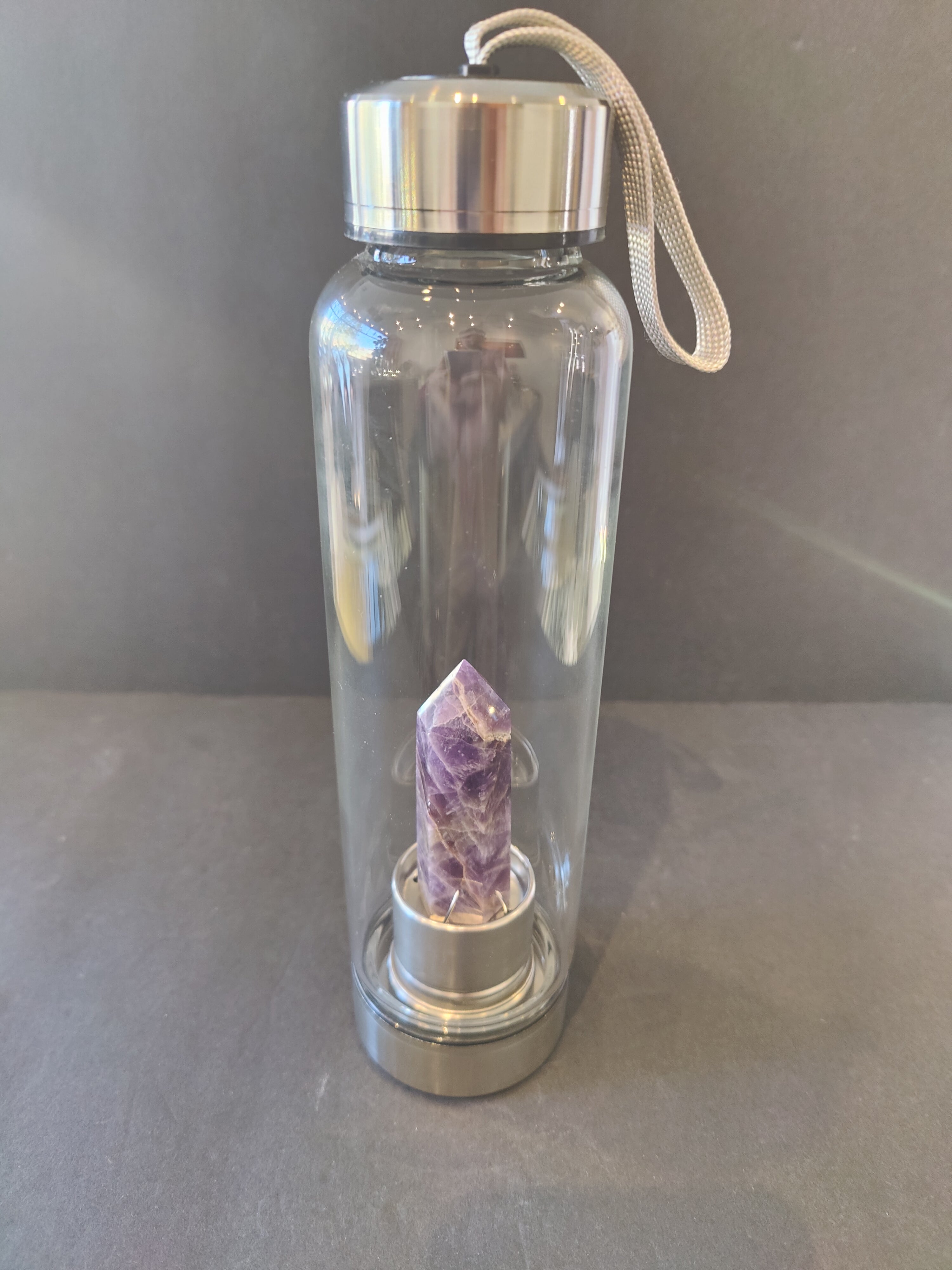Clear water bottle with a silver cap and a purple crystal inside on a gray surface.