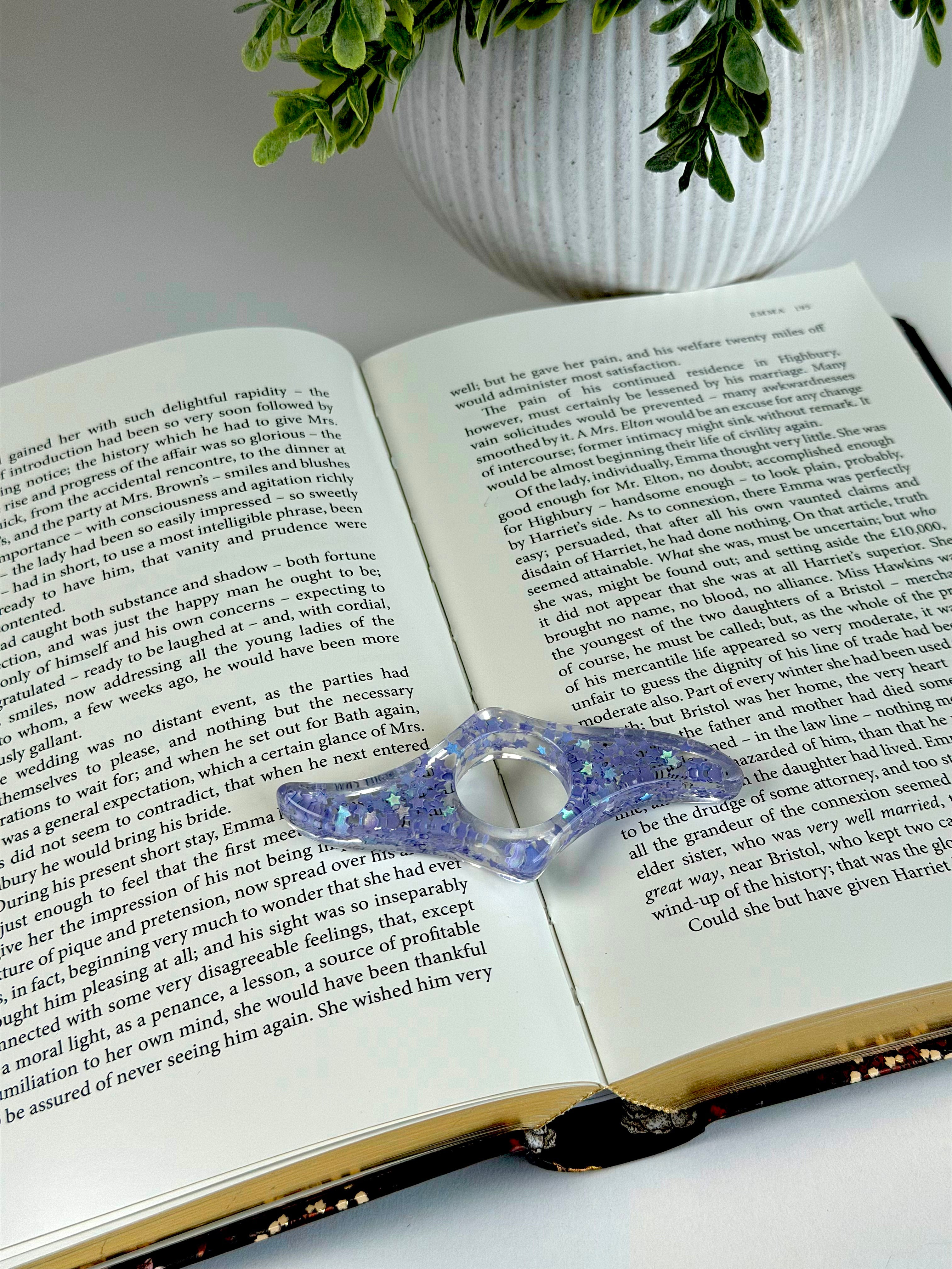 Open book with a decorative blue object on top, next to a white vase with greenery.