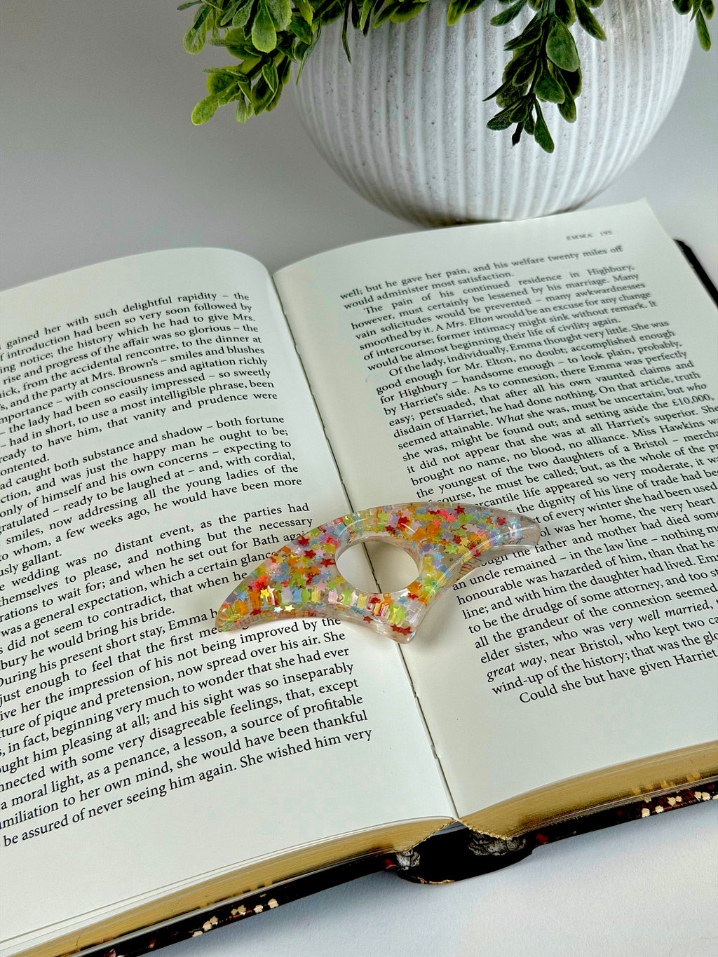 Open book with a colorful hair clip on a white surface, potted plant in the background