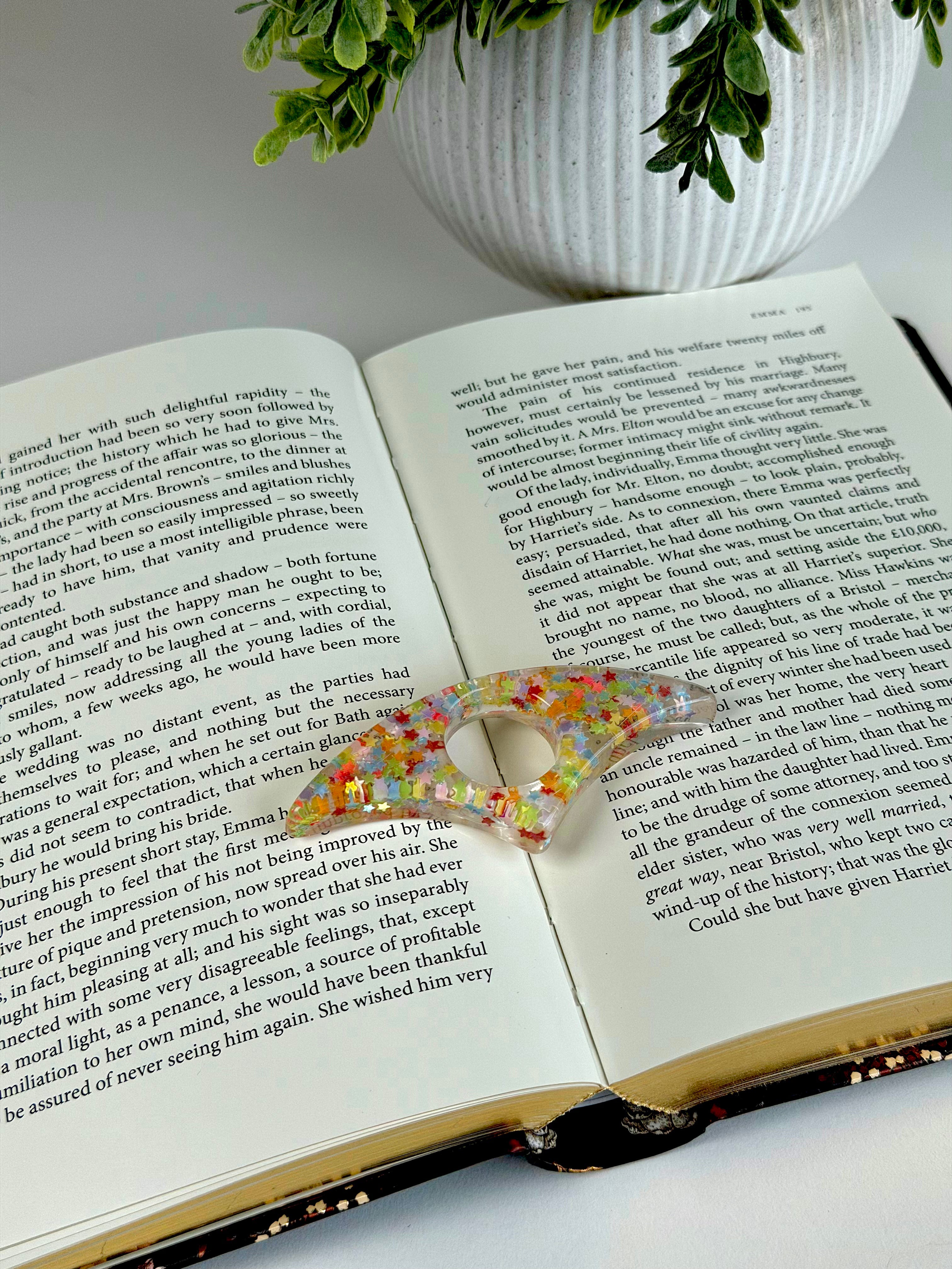 Open book with a colorful hair clip on a white surface, potted plant in the background