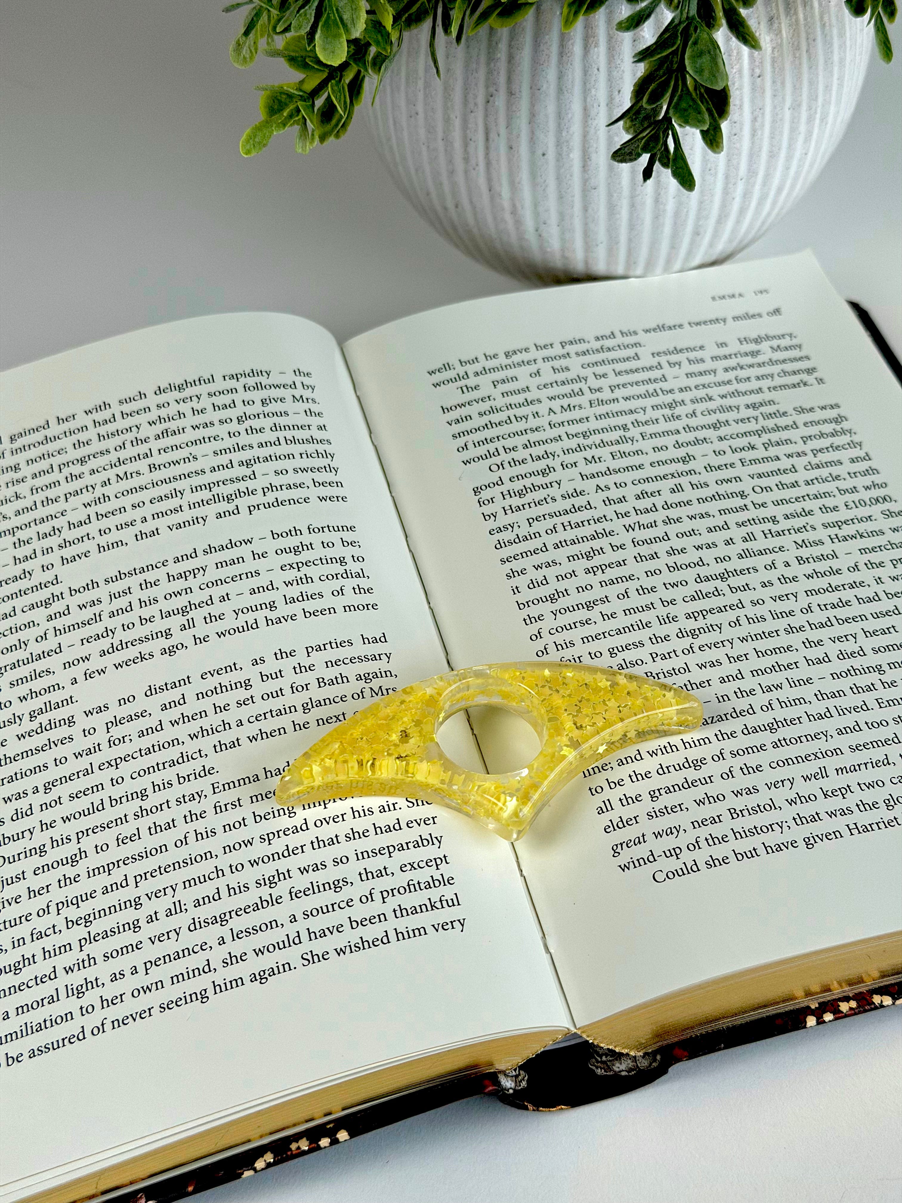 Open book with a yellow lace bookmark on a white surface, with a potted plant in the background.