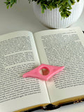 Open book with a pink page marker on a white surface, next to a potted plant.