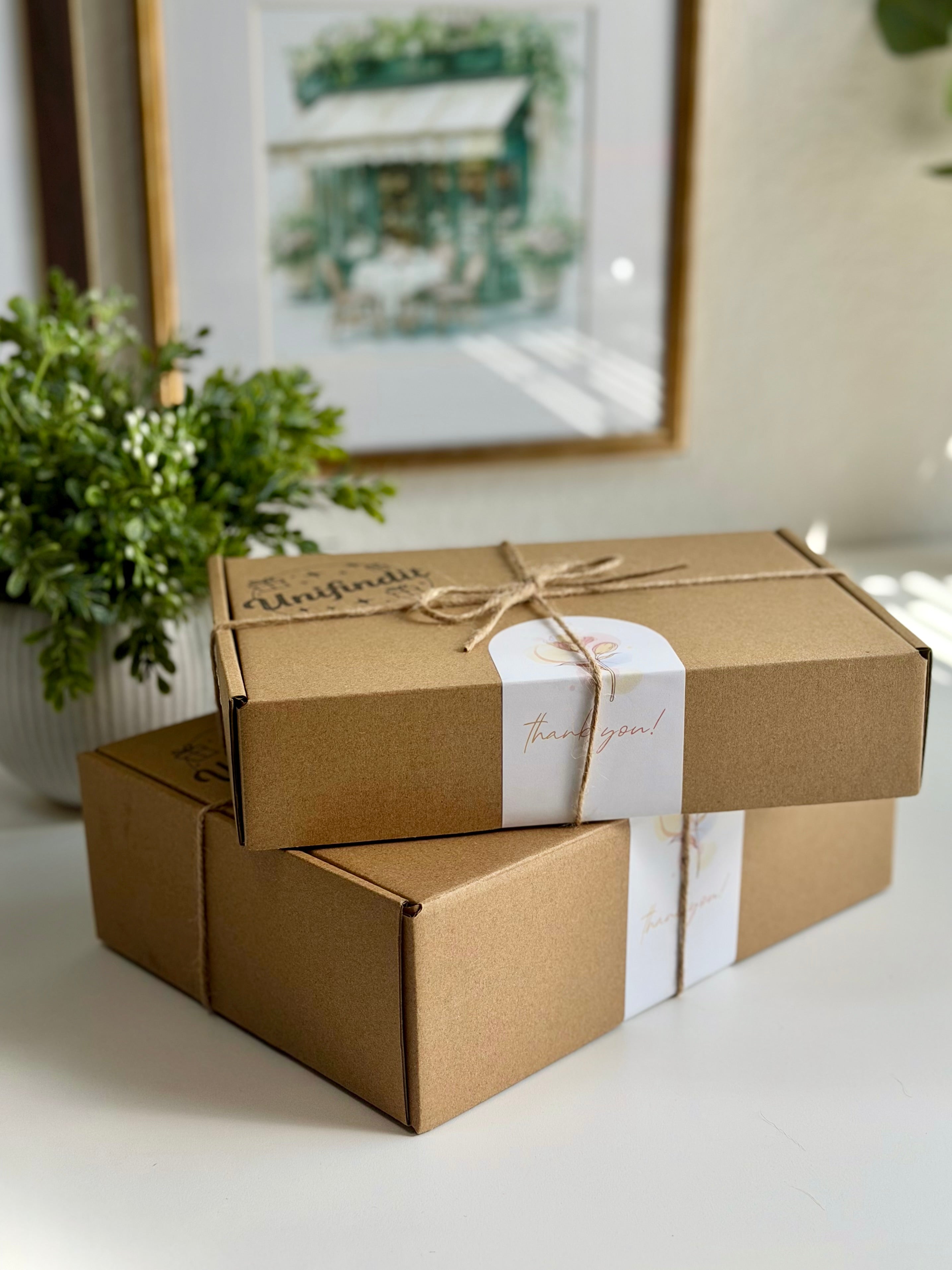 Two brown gift boxes with white ribbons on a light surface, with a plant and framed picture in the background.