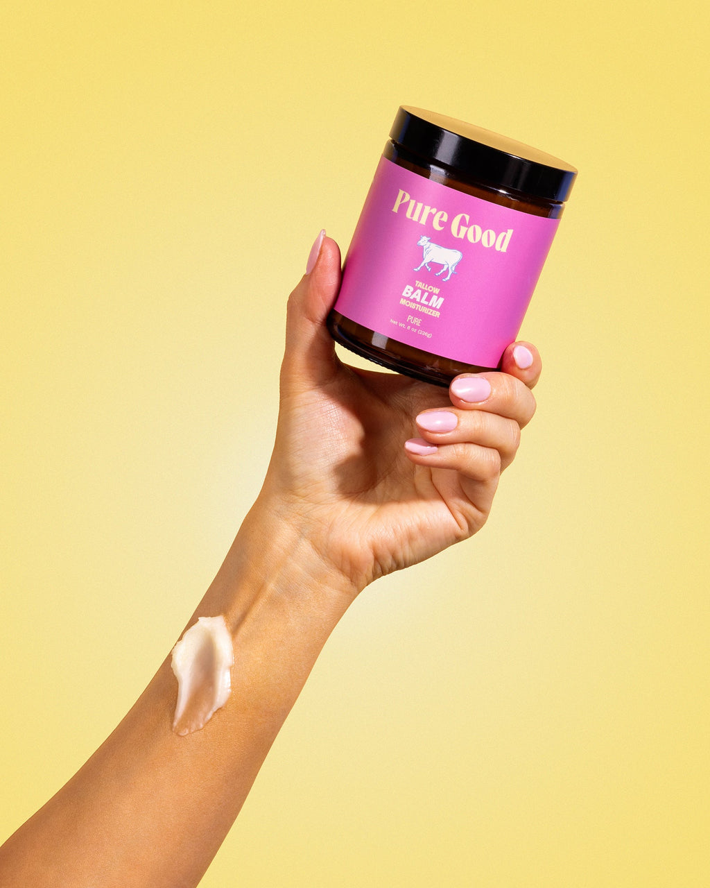 Hand holding a jar of 'Pure Good' balm with a swatch on a yellow background