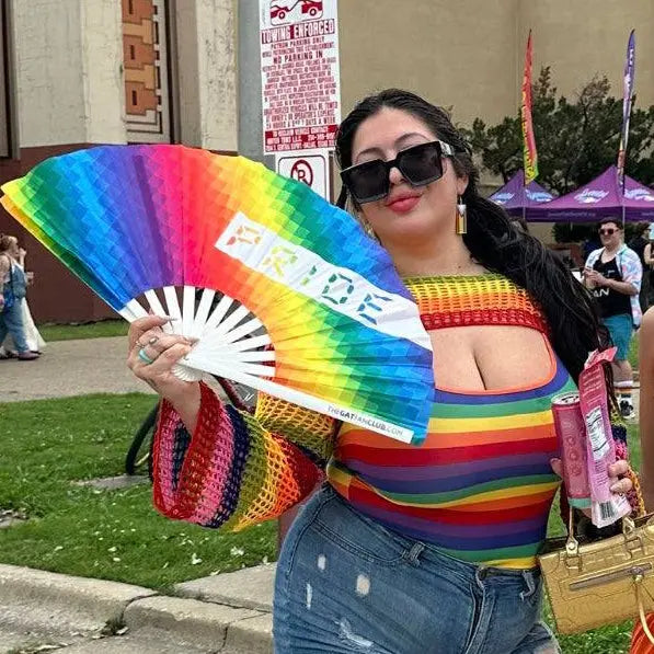 Person wearing a rainbow-colored top and holding a Pride fan outdoors.