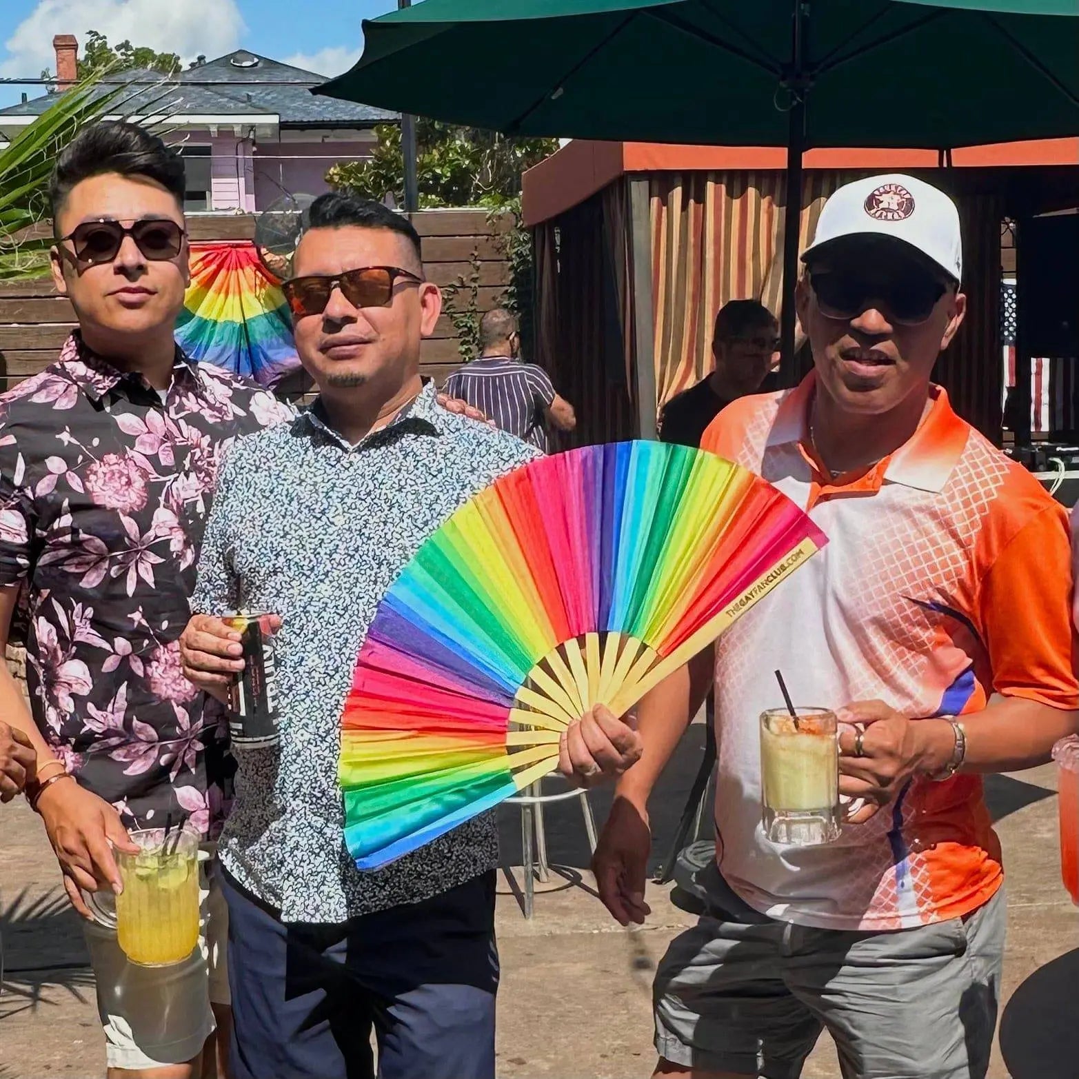 Three men standing outdoors, one holding a colorful fan, with a rainbow flag in the background.