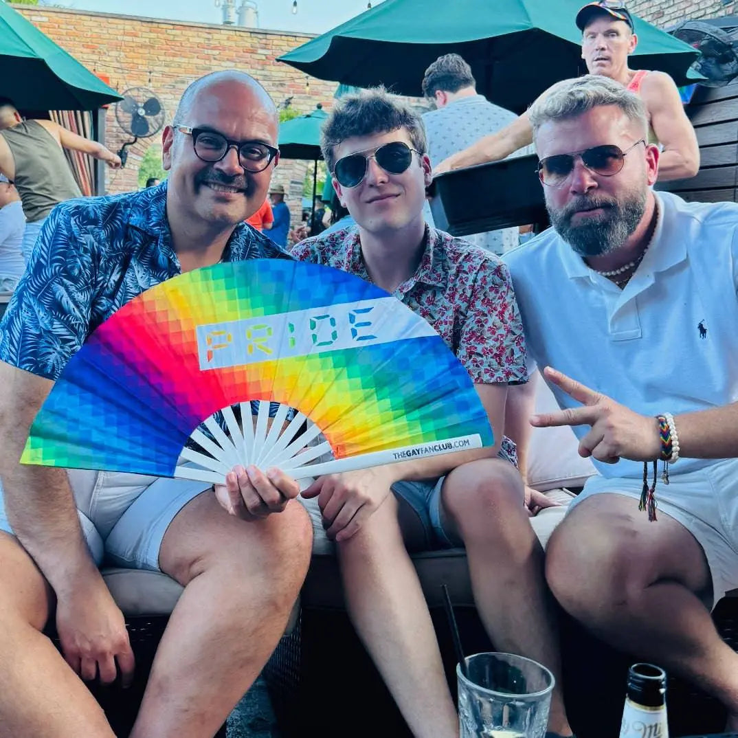 Three men sitting together with a rainbow 'PRIDE' fan at an outdoor event.