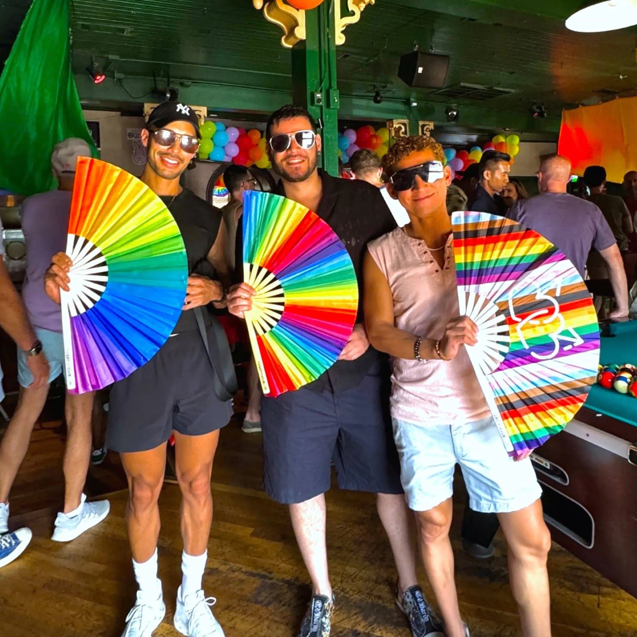 Three people holding rainbow-colored fans in a festive indoor setting with colorful decorations.