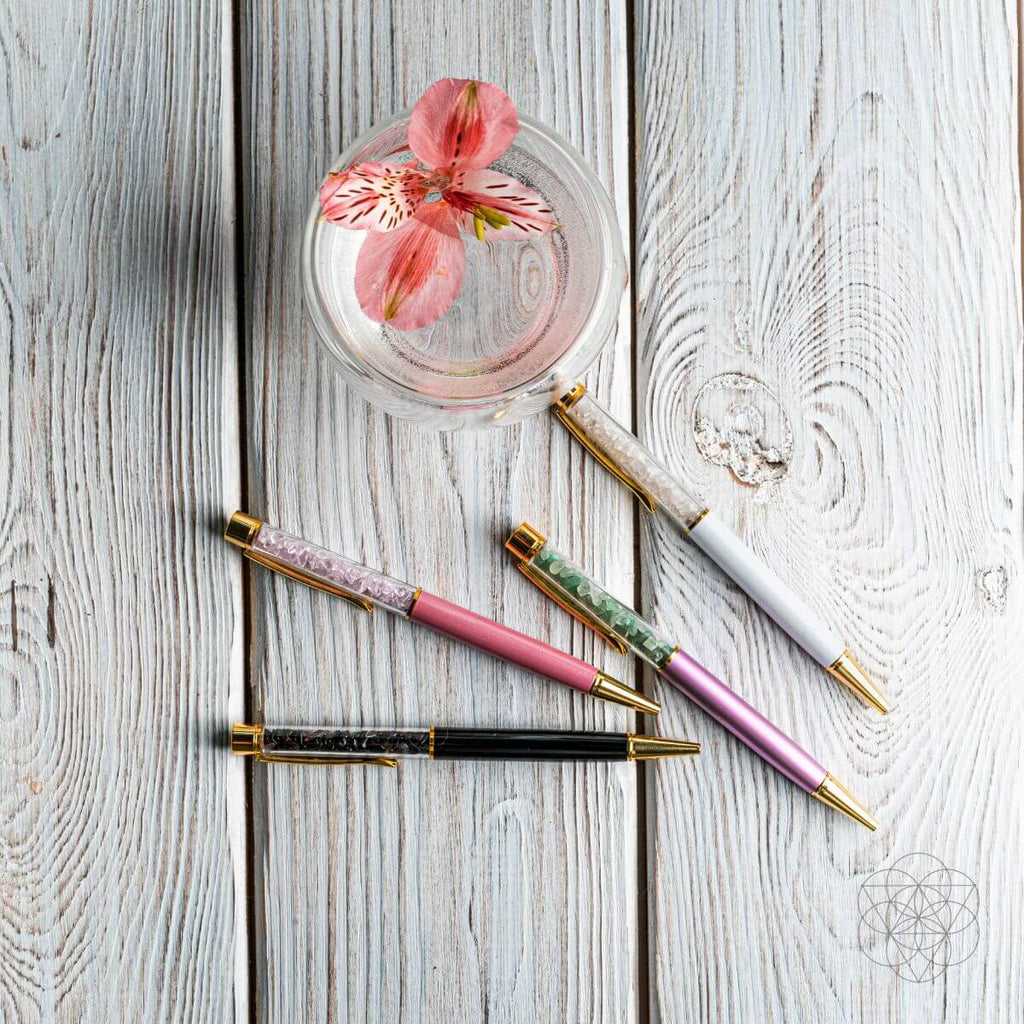Four decorative pens with gemstone accents on a wooden surface, one with a pink flower in a glass.