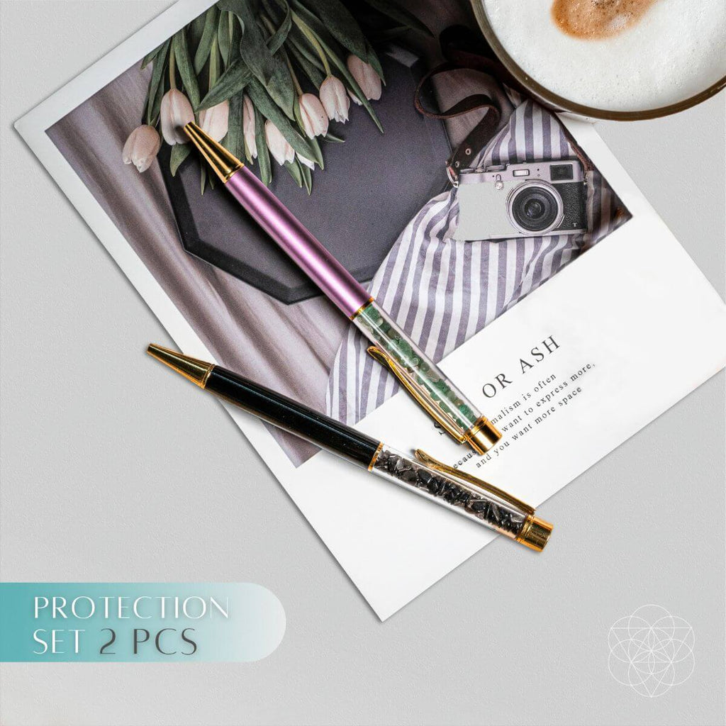 Two pens on a surface with a photo, camera, and coffee cup in the background.
